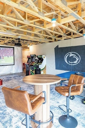 A room with a wooden ceiling, a round table, two bar stools, a projector screen, and arcade games. - Welcome to Happy Valley Retreat! Features a golf simulator and a game room! (Bellefonte)