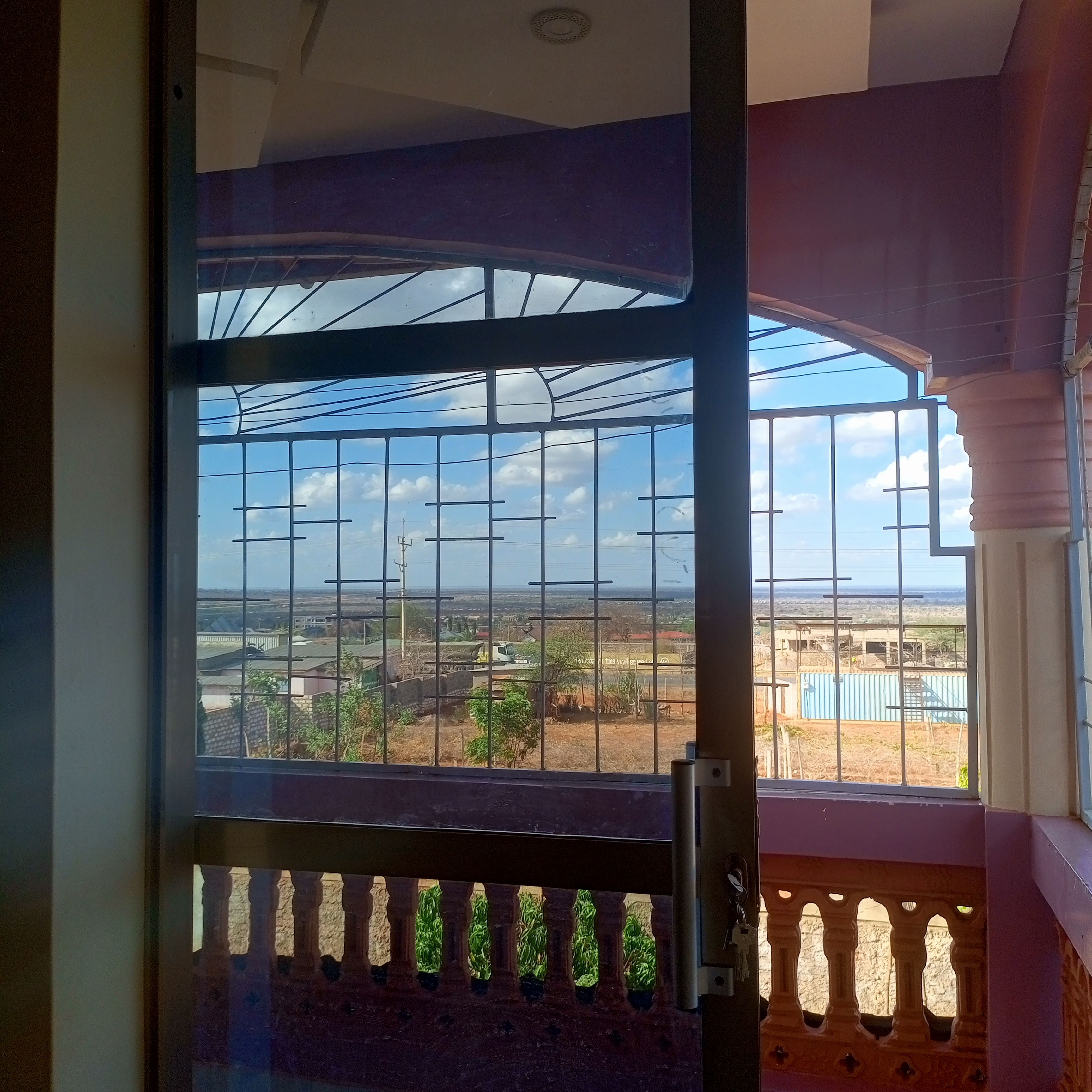 A room with a large window featuring a metal grid, a balcony with a railing, and a view of a landscape with buildings and vegetation.