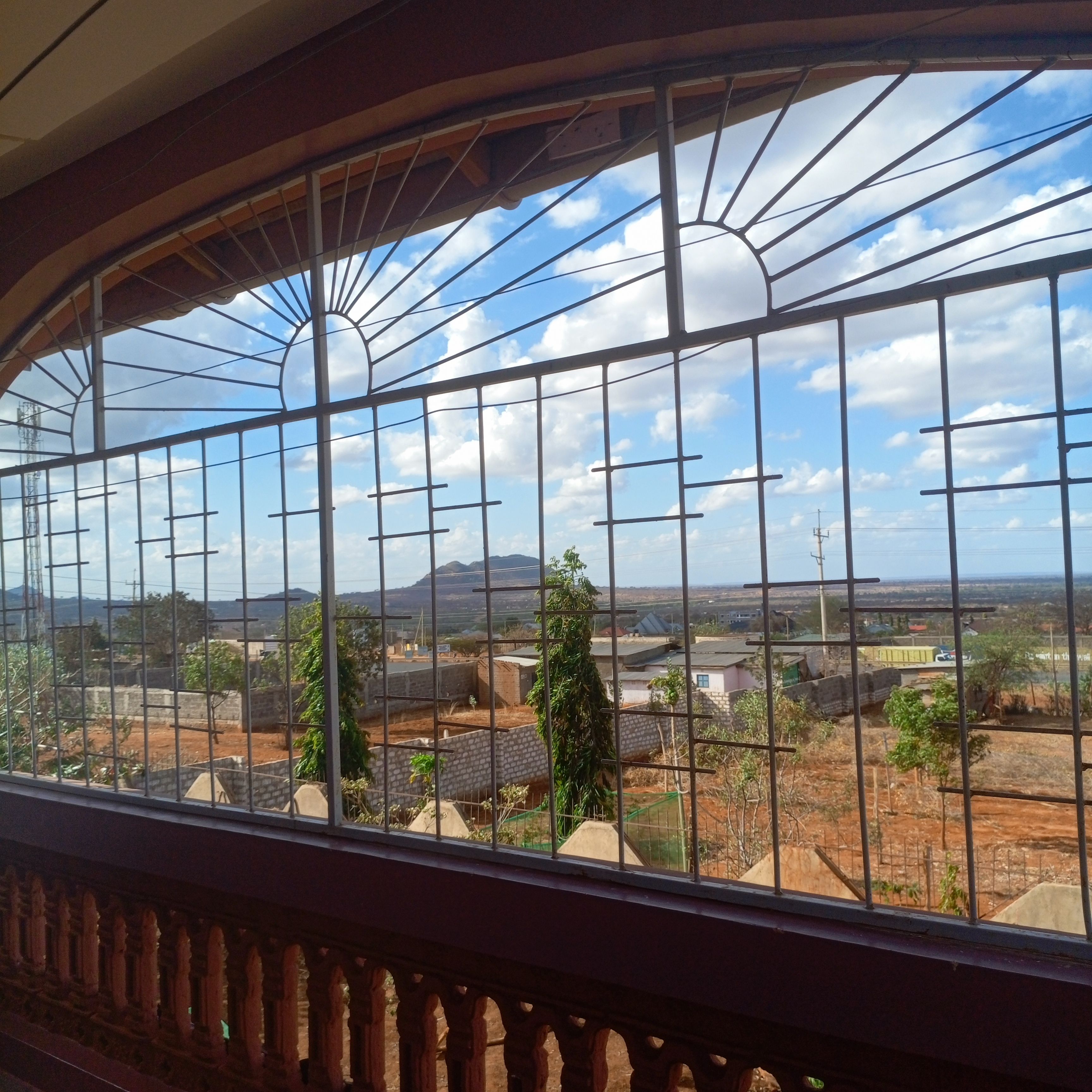 A view through a window with metal bars, featuring a landscape with buildings, trees, and a clear sky.