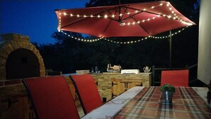 Un área de comedor al aire libre con un paraguas rojo, un horno de ladrillo y una mesa con mantel a cuadros.