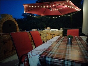 Een terras met een rode parasol, een bakoven en een tafel met een geruit tafelkleed.