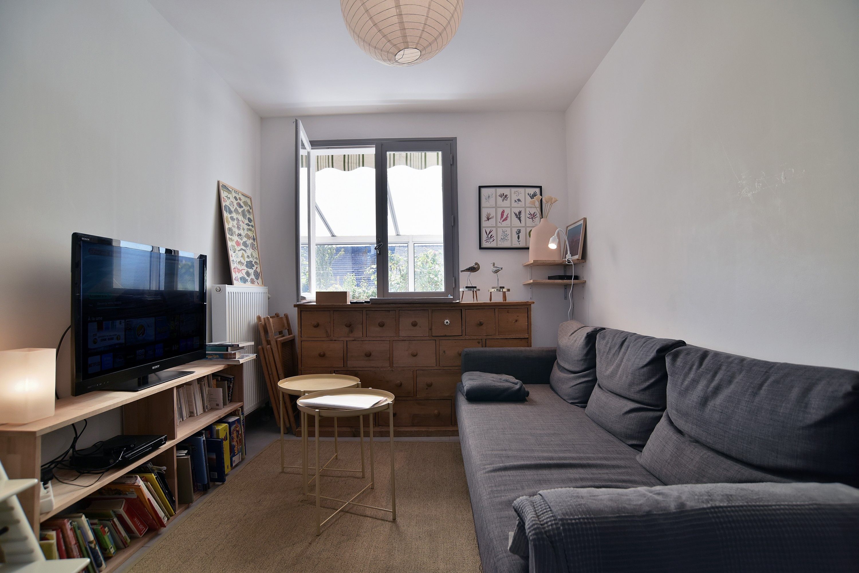 A living room with a grey sofa, a television, a wooden cabinet, and a window with blinds.