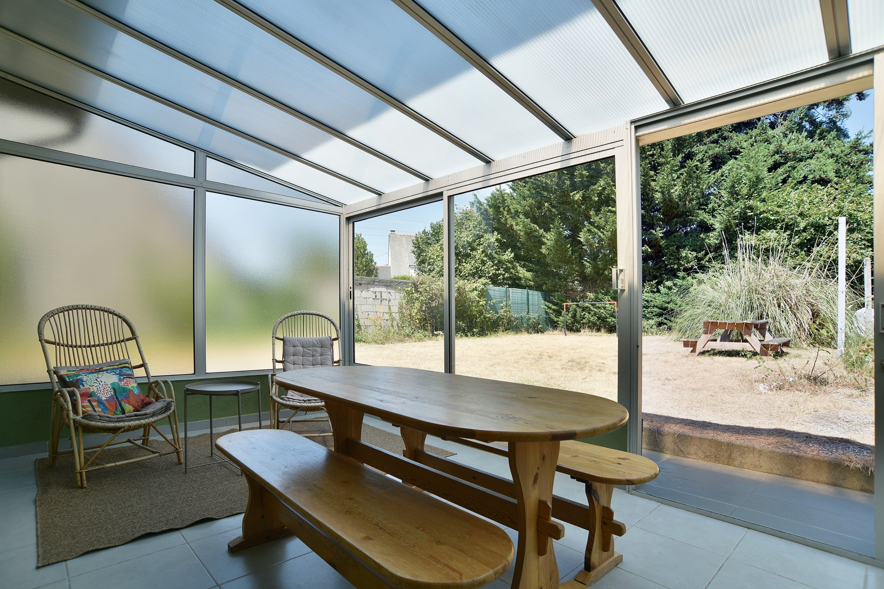 A covered patio with a wooden table and chairs, large windows, and a view of the outdoors.