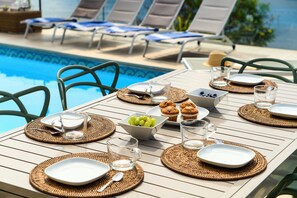 Zona de comedor junto a la piscina con sillas de descanso, una mesa para dos con platos, vasos, un cuenco de uvas y muffins.