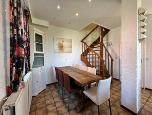 A dining area with a wooden table and chairs, a white radiator, a window with curtains, a staircase, and a wall-mounted artwork. - Comfortable Heuvelland Family Escape (Gulpen)