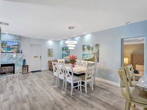 A dining area with a wooden floor, white chairs, a central table with a vase of flowers, and wall art depicting tropical scenes. - 3 min walk2 bch•Heat Pool•Bbq•100”TV•On Ocean Blvd (Fort Lauderdale)