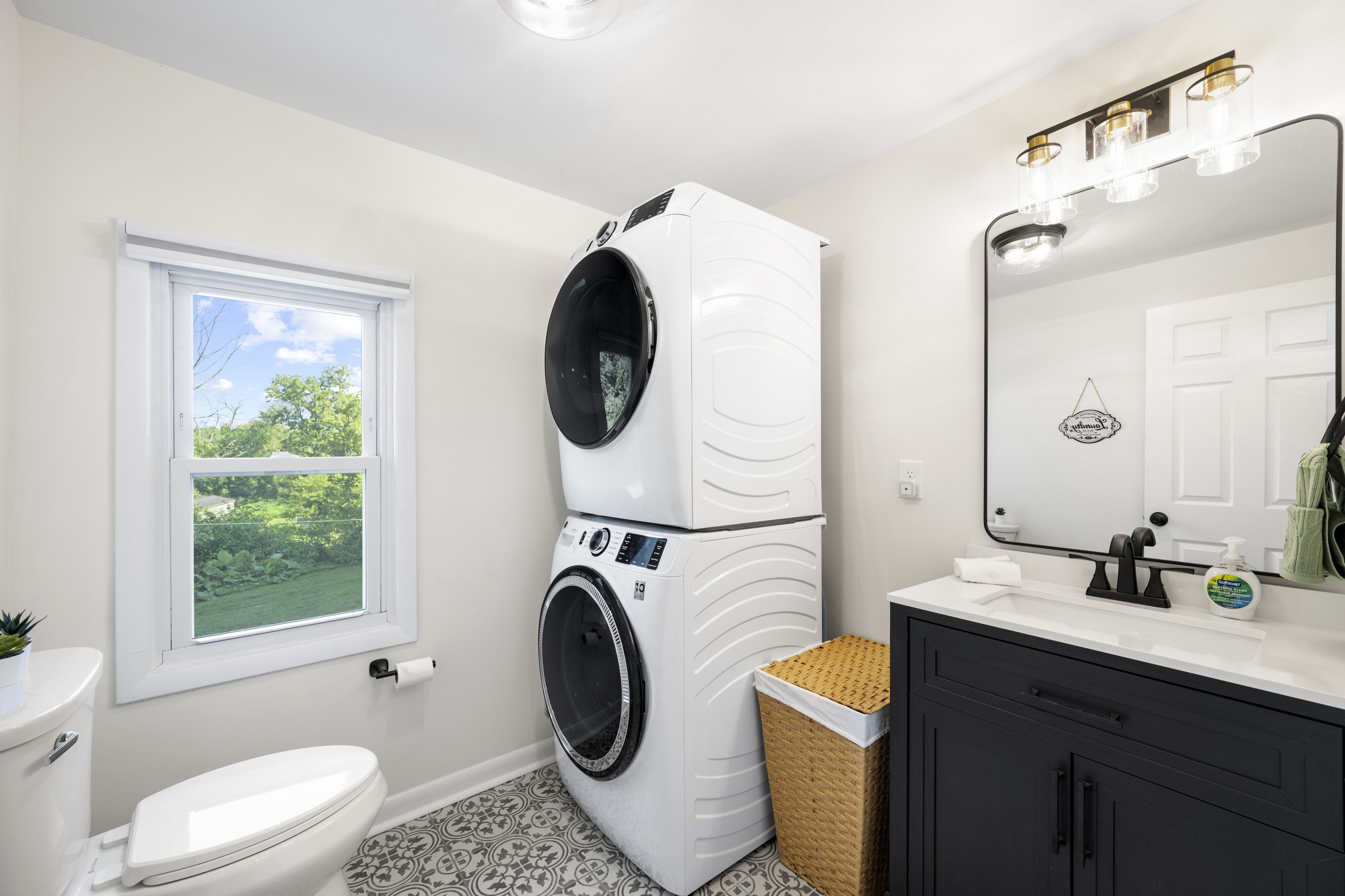 A compact bathroom with a white toilet, a washing machine, a sink with a mirror, and a small window.