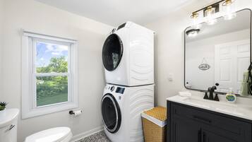 A compact bathroom with a white toilet, a washing machine, a sink with a mirror, and a small window.