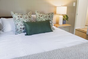 Un lit bien fait, avec une literie blanche et des coussins décoratifs, une lampe de chevet et une petite plante en pot posée sur une table de chevet.