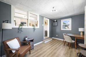 Une pièce avec un plancher en bois, un fauteuil en cuir marron, une table à manger avec des chaises, un petit bureau et une fenêtre donnant sur l’extérieur.