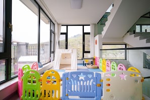A room with large windows, a play area with colorful plastic play structures, and a white tent-like playhouse.