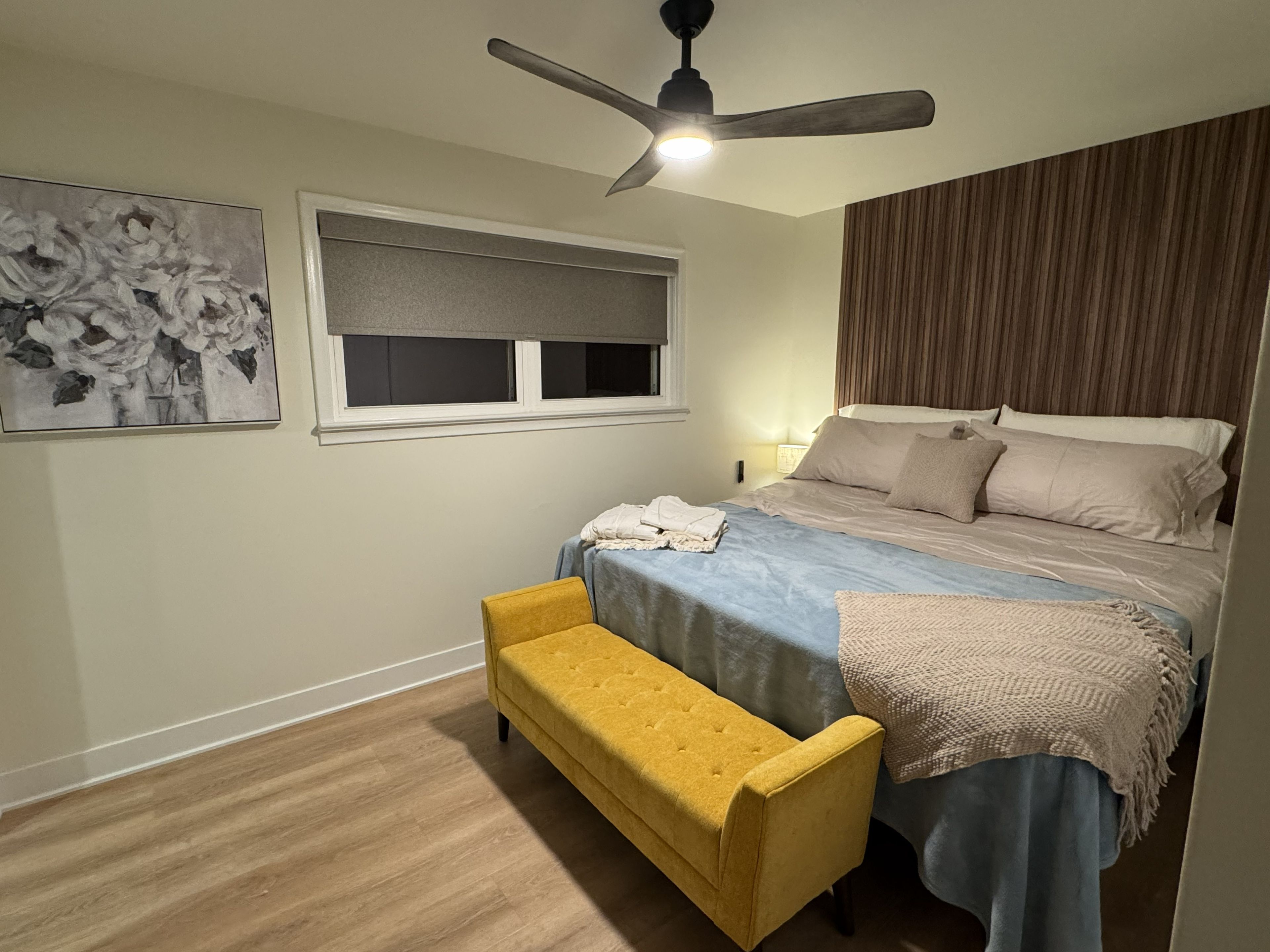 Quarto com cama, um banco, ventoinha no teto, janela com persianas e um quadro na parede.