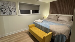 Un dormitorio con una cama, un banco, un ventilador de techo, una ventana con persianas y un cuadro en la pared.