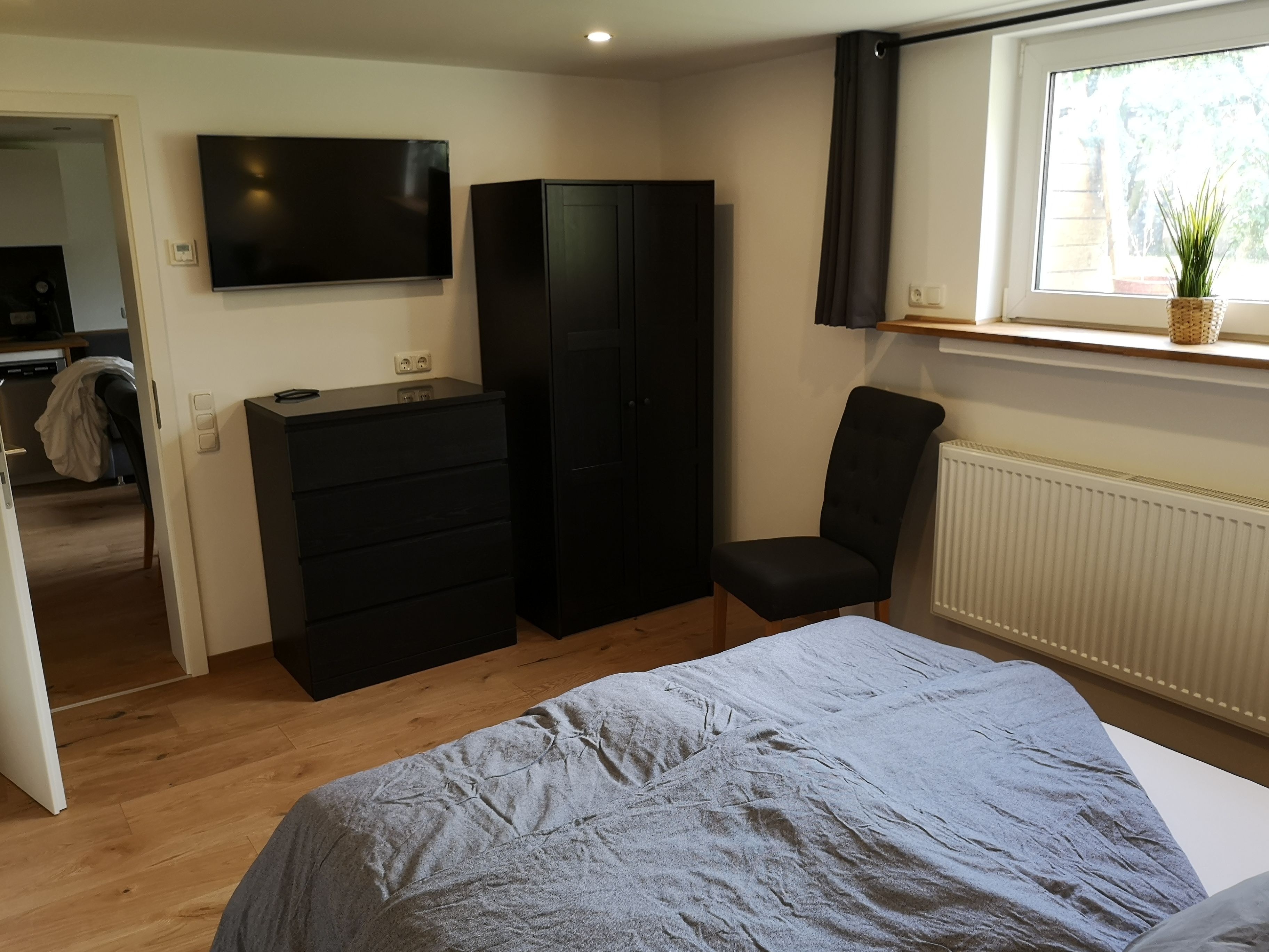 Een slaapkamer met een bed, een kapstok, een stoel, een kledingkast, een televisie en een raam met een plant.