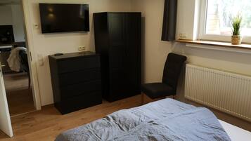 Een slaapkamer met een bed, een kapstok, een stoel, een kledingkast, een televisie en een raam met een plant.