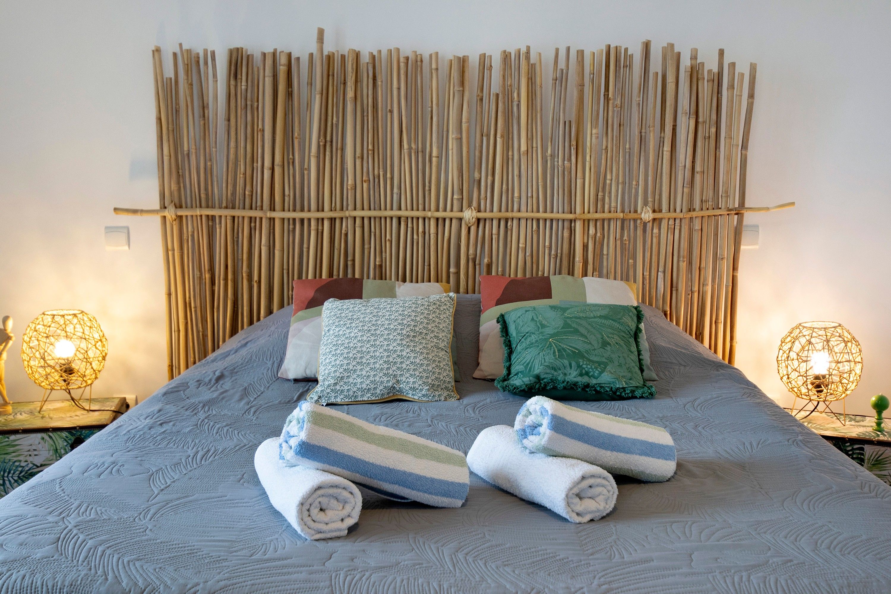 A bed with a grey quilt, several pillows, and rolled towels.