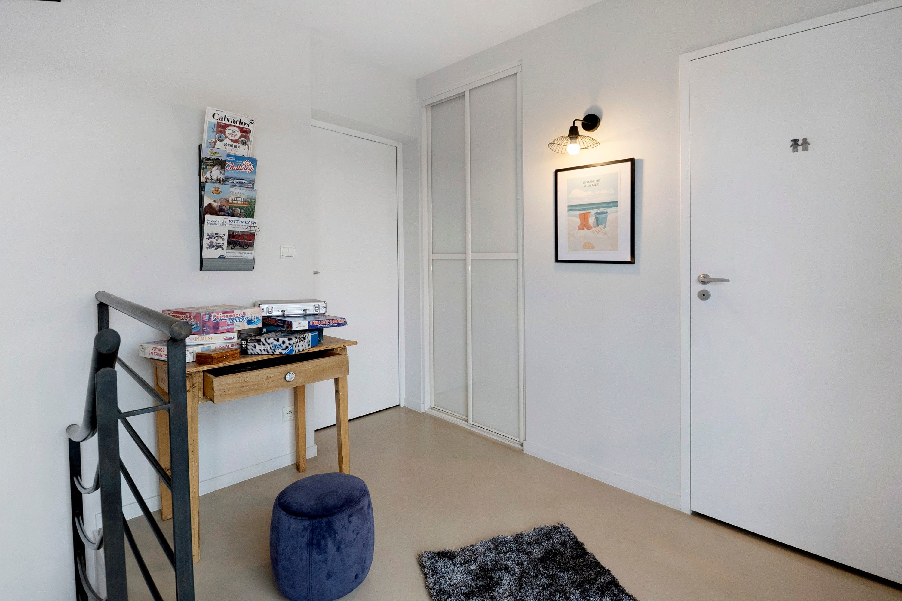 Un pasillo con una mesa de madera, un otoman azul, un cuadro enmarcado y una puerta blanca.