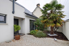 A residential building with a white exterior, a sloped roof, and a small garden area with gravel, a palm tree, and a variety of shrubs.