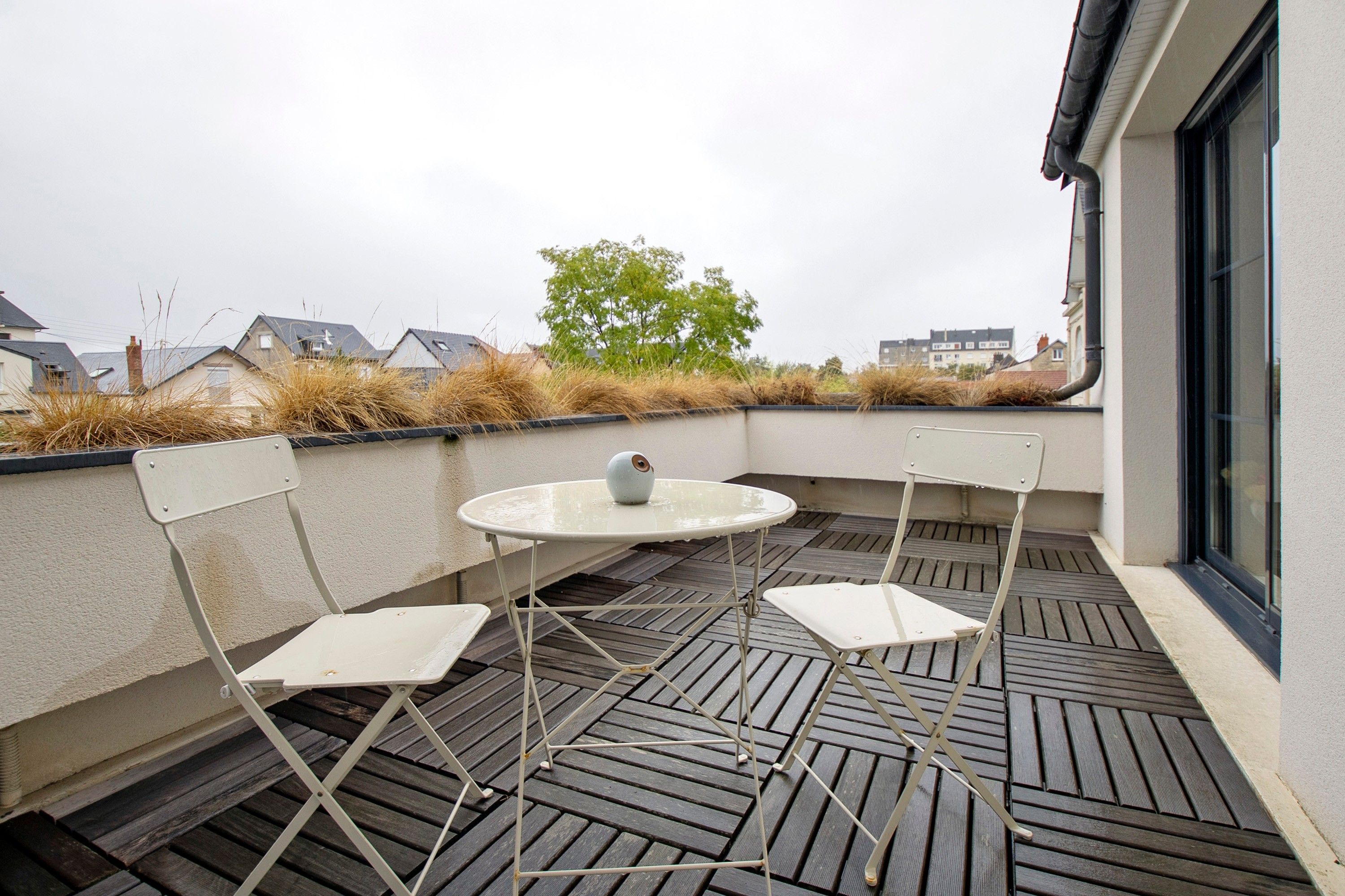 Un petit espace extérieur aménagé avec une table ronde et deux chaises, un pot de fleurs et une vue sur le quartier.