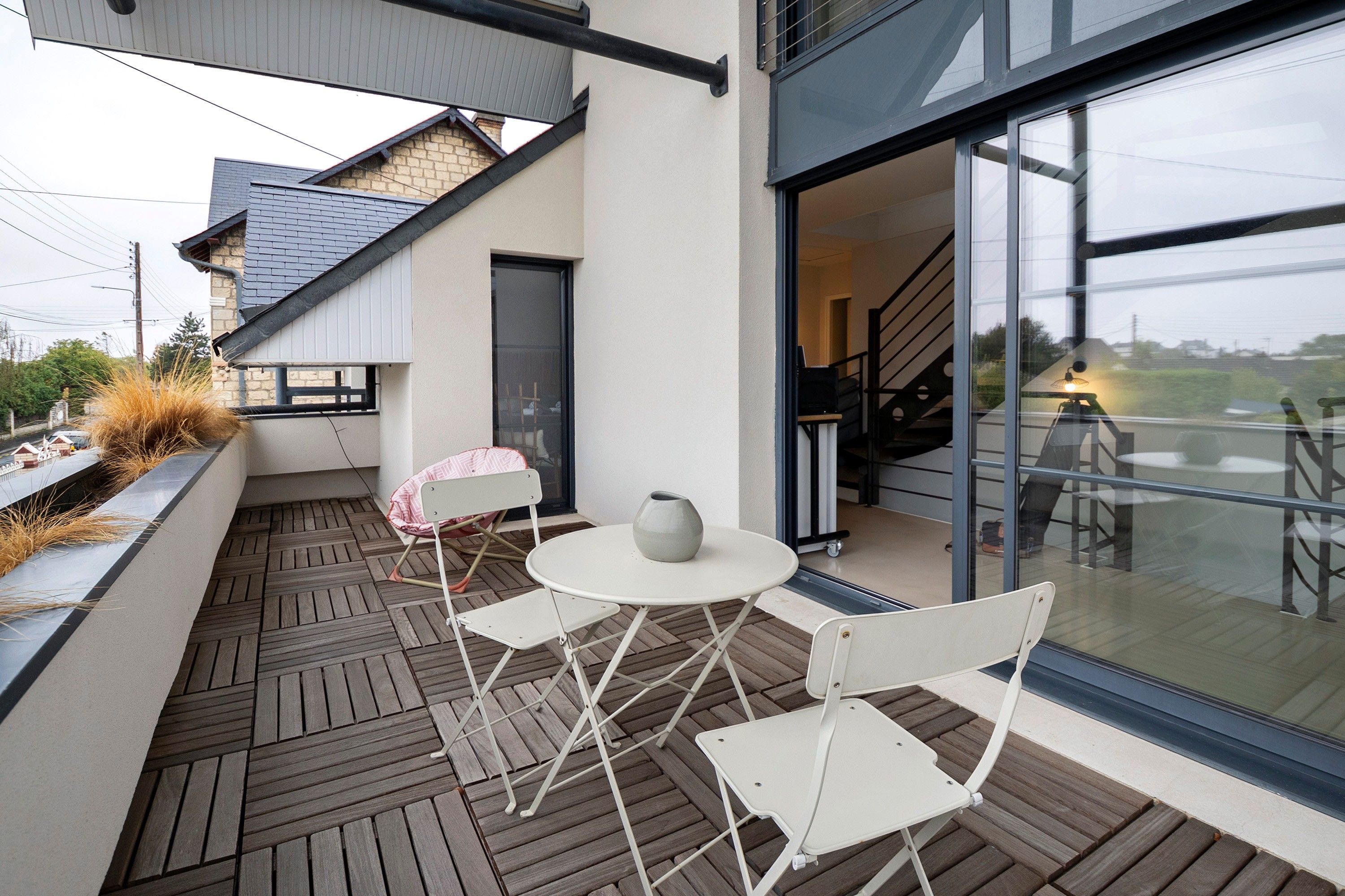Um pátio externo com mesa e cadeiras, um vaso de planta e vista do interior através de uma porta de vidro.