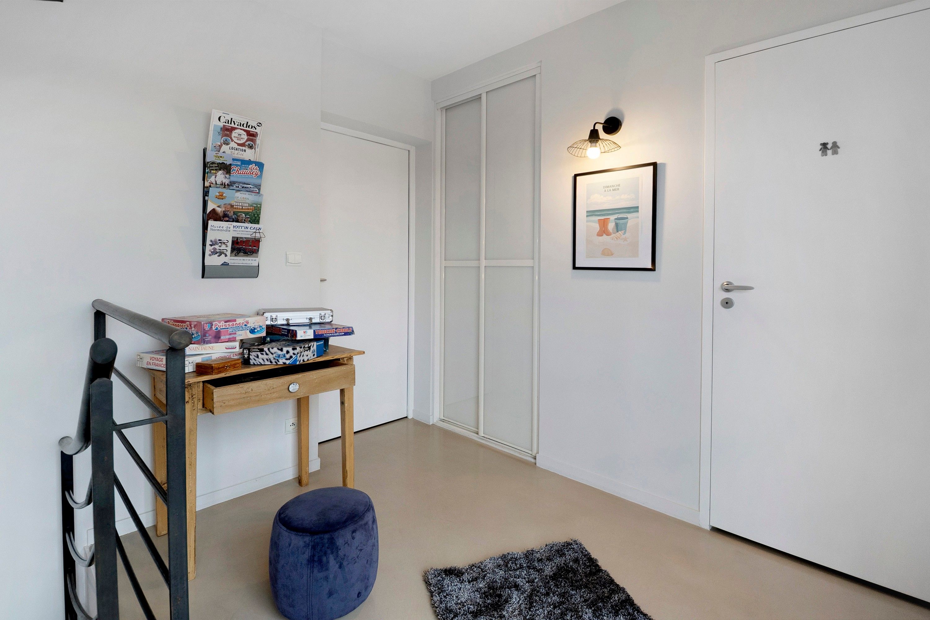 Un pasillo con una mesa de madera que sostiene juegos de mesa, un otoman azul, un cuadro en la pared y una puerta blanca.