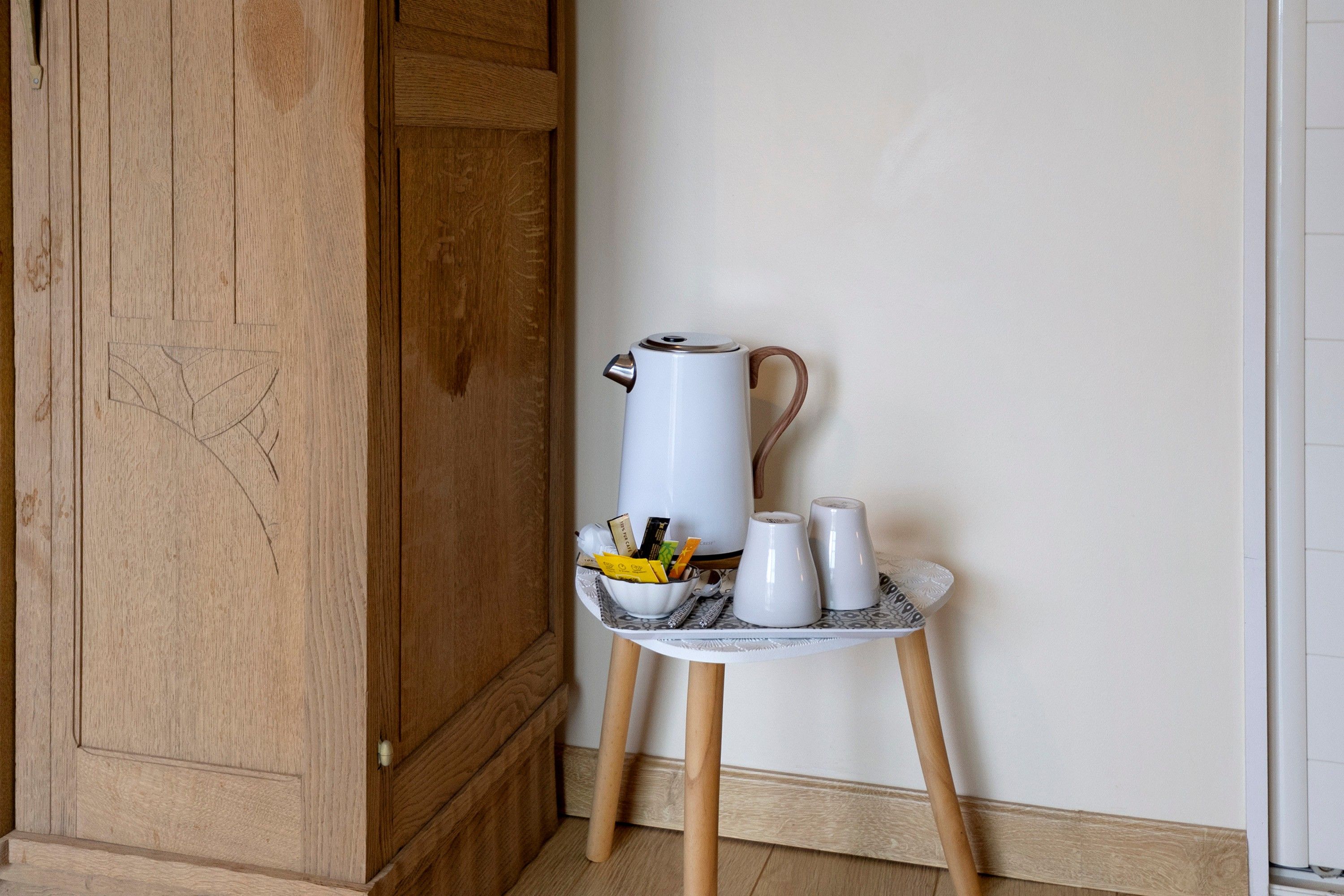 Una tetera blanca sobre un soporte de madera, junto a tazas y un cuenco pequeño con bolsitas de té y una cuchara.