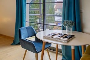 A room with a round table, a blue chair, a vase with flowers, and a view of a balcony with greenery.