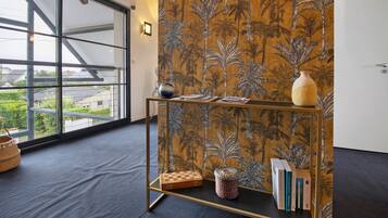 Ein Zimmer mit einem großen Fenster, einer Wand mit tropischem Muster, einem kleinen Tisch mit Büchern und Schachbrett sowie einer Vase auf dem Tisch.