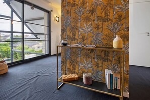 Una habitación con un ventanal grande, una pared con motivo tropical, una mesita con libros y un tablero de ajedrez, y un florero sobre la mesita.