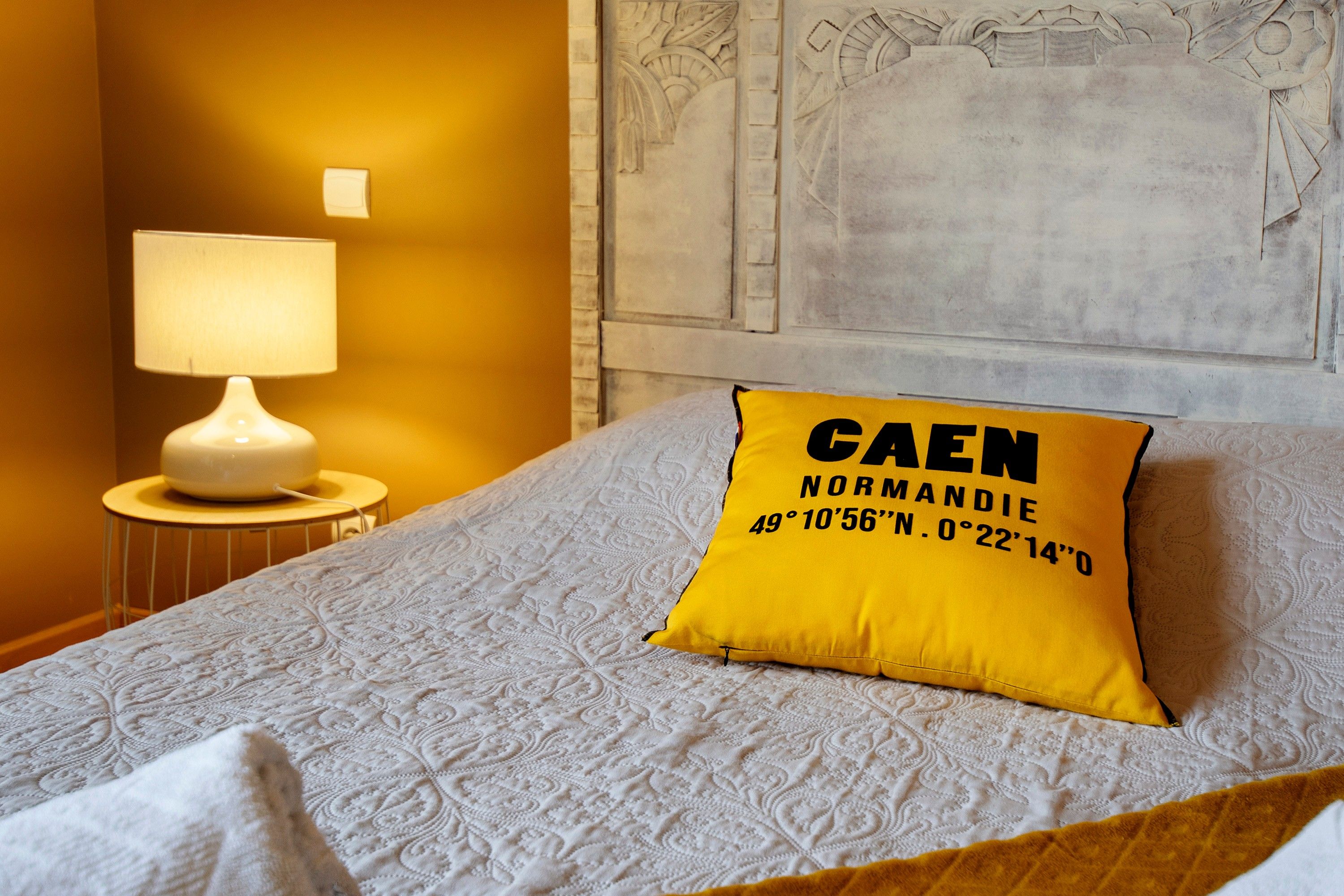 A bed with a patterned bedspread, a yellow pillow with text, a bedside lamp, and a wall with carved designs.