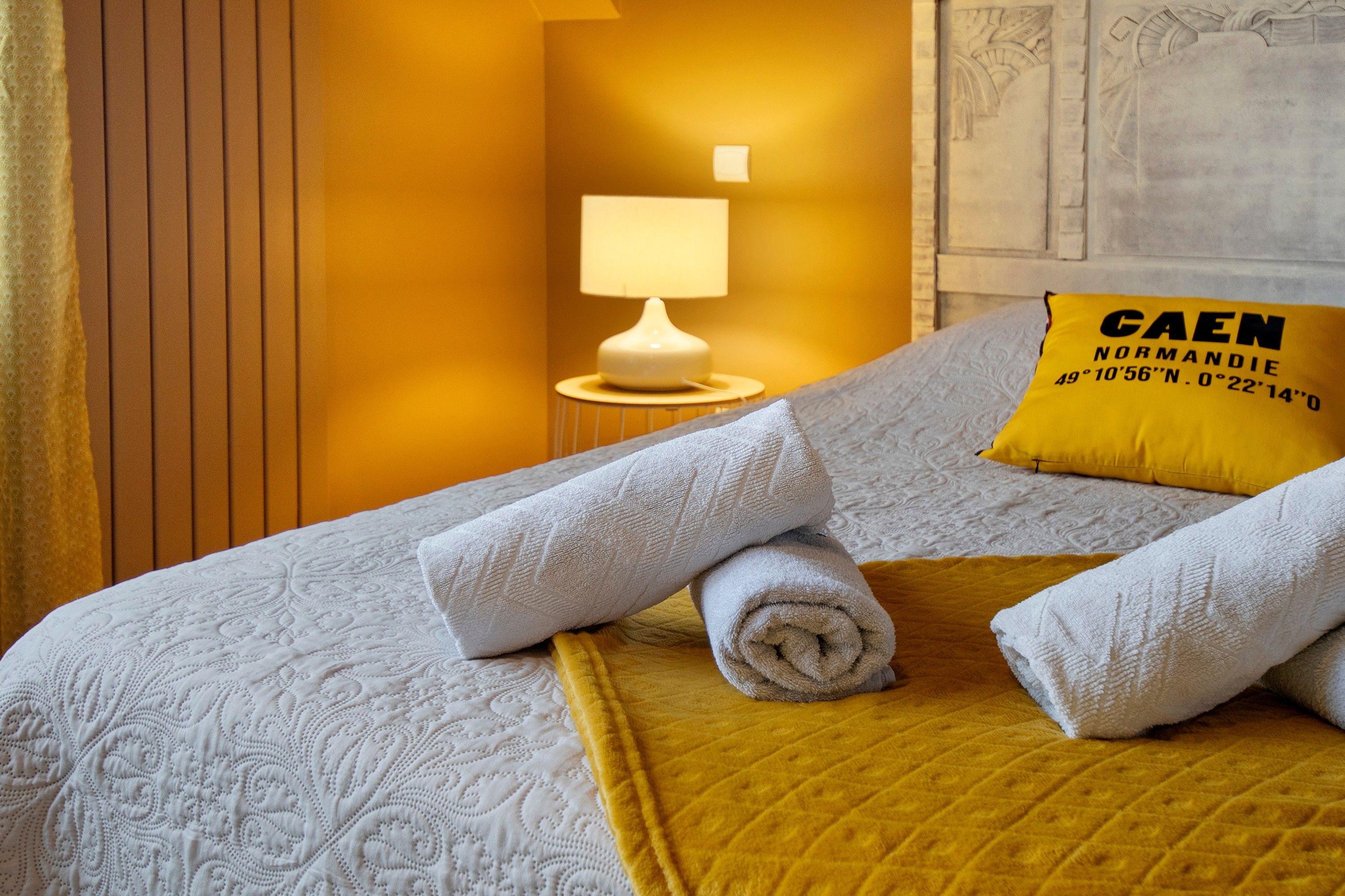 Cama bem arrumada com toalhas enroladas, um cobertor amarelo e um travesseiro com texto.