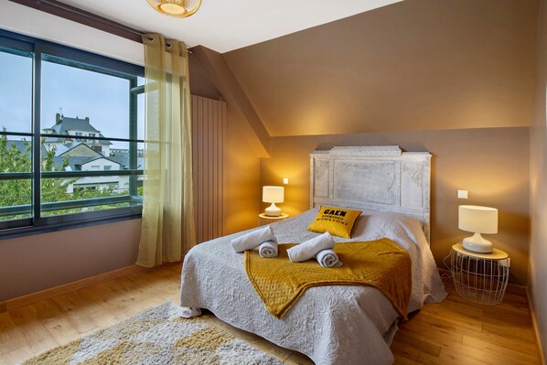 A hotel room with a bed, two bedside lamps, a window with curtains, a view of buildings outside, and a headboard with a plaque. - Bed and Breakfast 'Chambre Deuxième Étage' with Shared Terrace, Balcony and Wi-Fi (Caen)