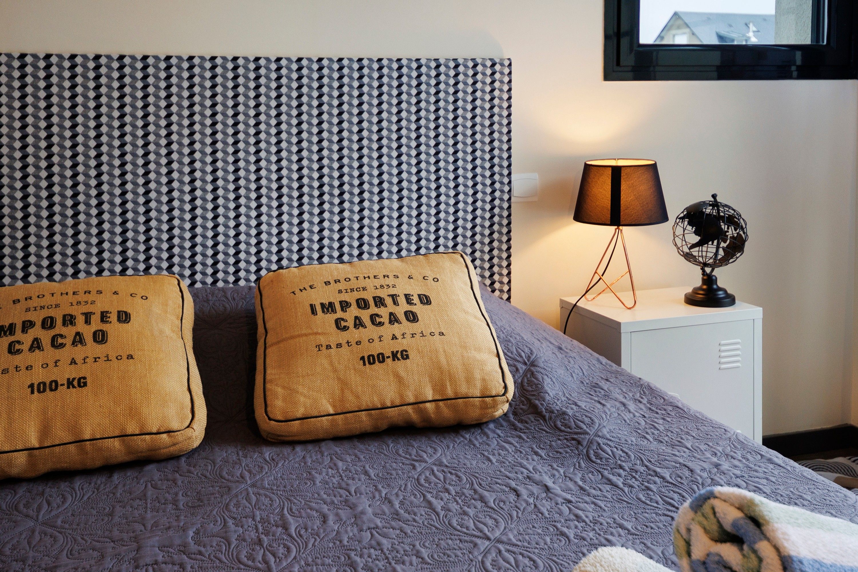 Un lit avec une tête de lit à motifs, deux oreillers portant l’inscription « Cacao importé, 100 kg », une lampe de chevet et un globe terrestre sur une table de chevet.