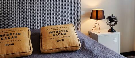Un llit amb un panell de capçal estampat, dos almoines amb l'etiqueta 'Imported Cacao 100-KG', una làmpada de nit i un glob dins d'una taula de nit.