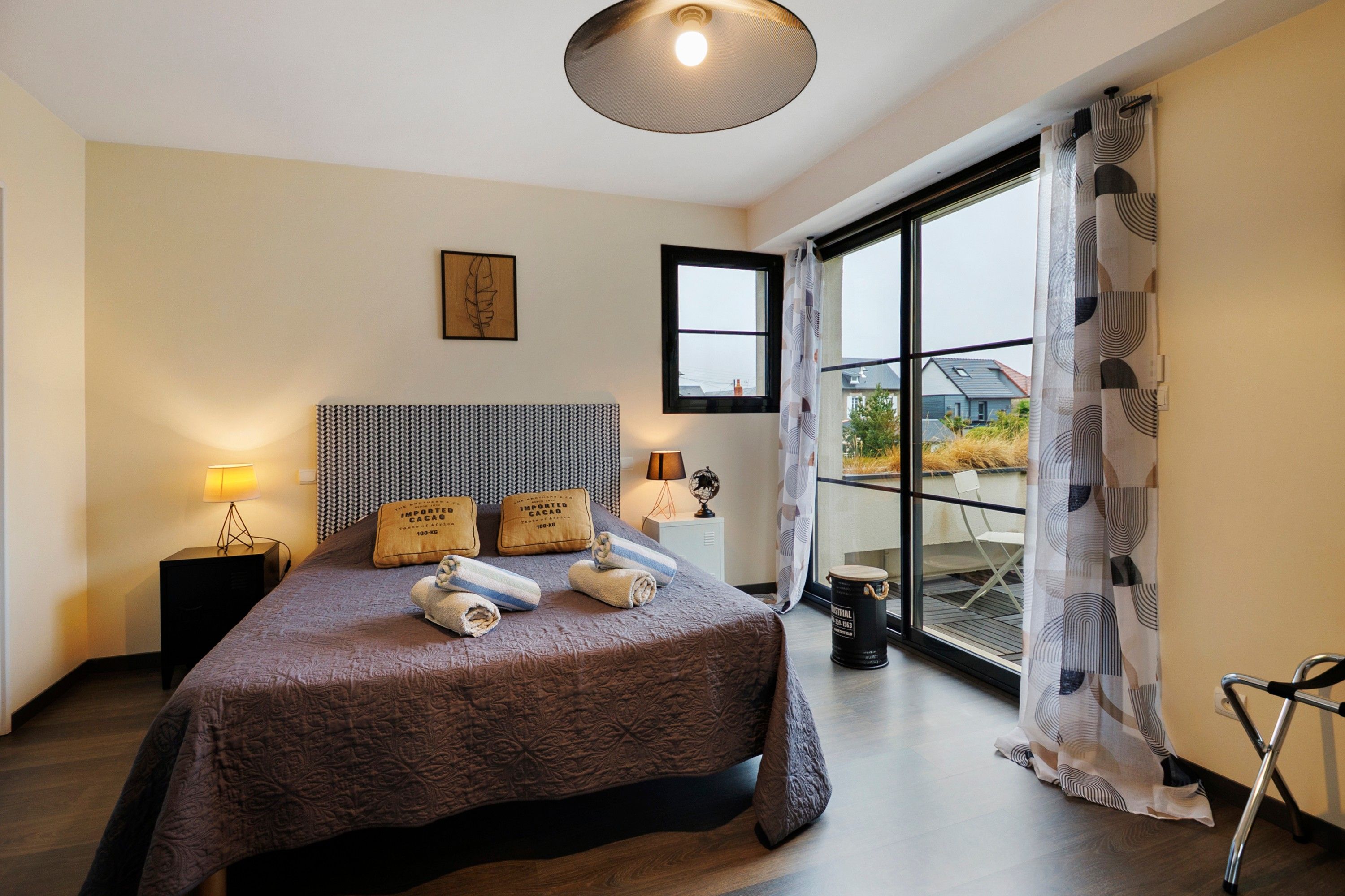 A modern bedroom with a large bed, a bedside table with a lamp, a window with curtains, and a view of the outside.