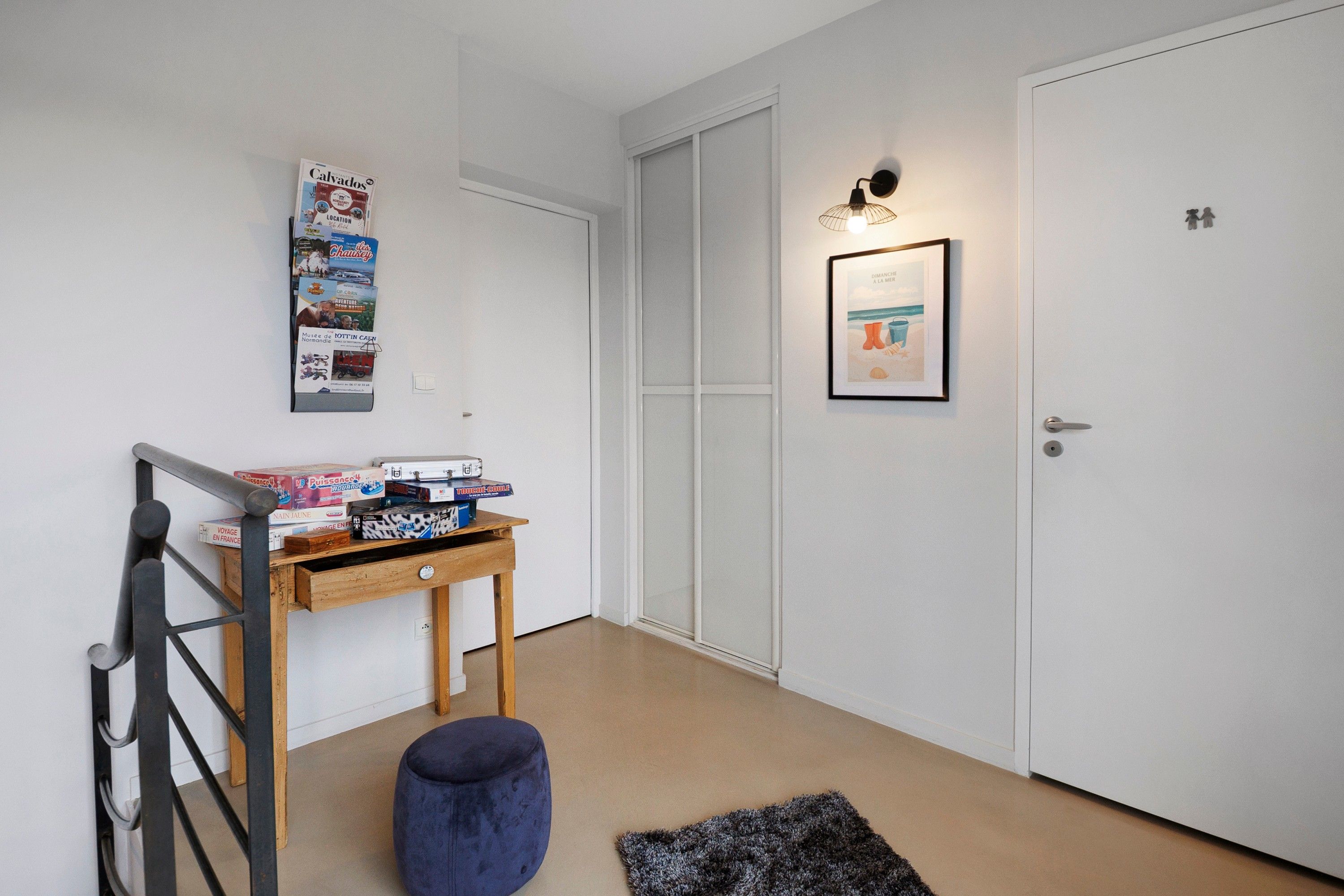 Un couloir avec une porte blanche, une petite table sur laquelle sont posés des jeux de société, un tableau encadré accroché au mur et un pouf bleu.