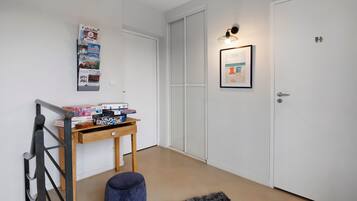 Een gang met een witte deur, een kleine tafel met gezelschapsspellen, een lijst met een foto aan de muur en een blauwe poef.