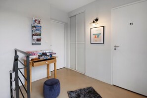 Un pasillo con una puerta blanca, una pequeña mesa con juegos de mesa, un cuadro en la pared y un otoman azul.