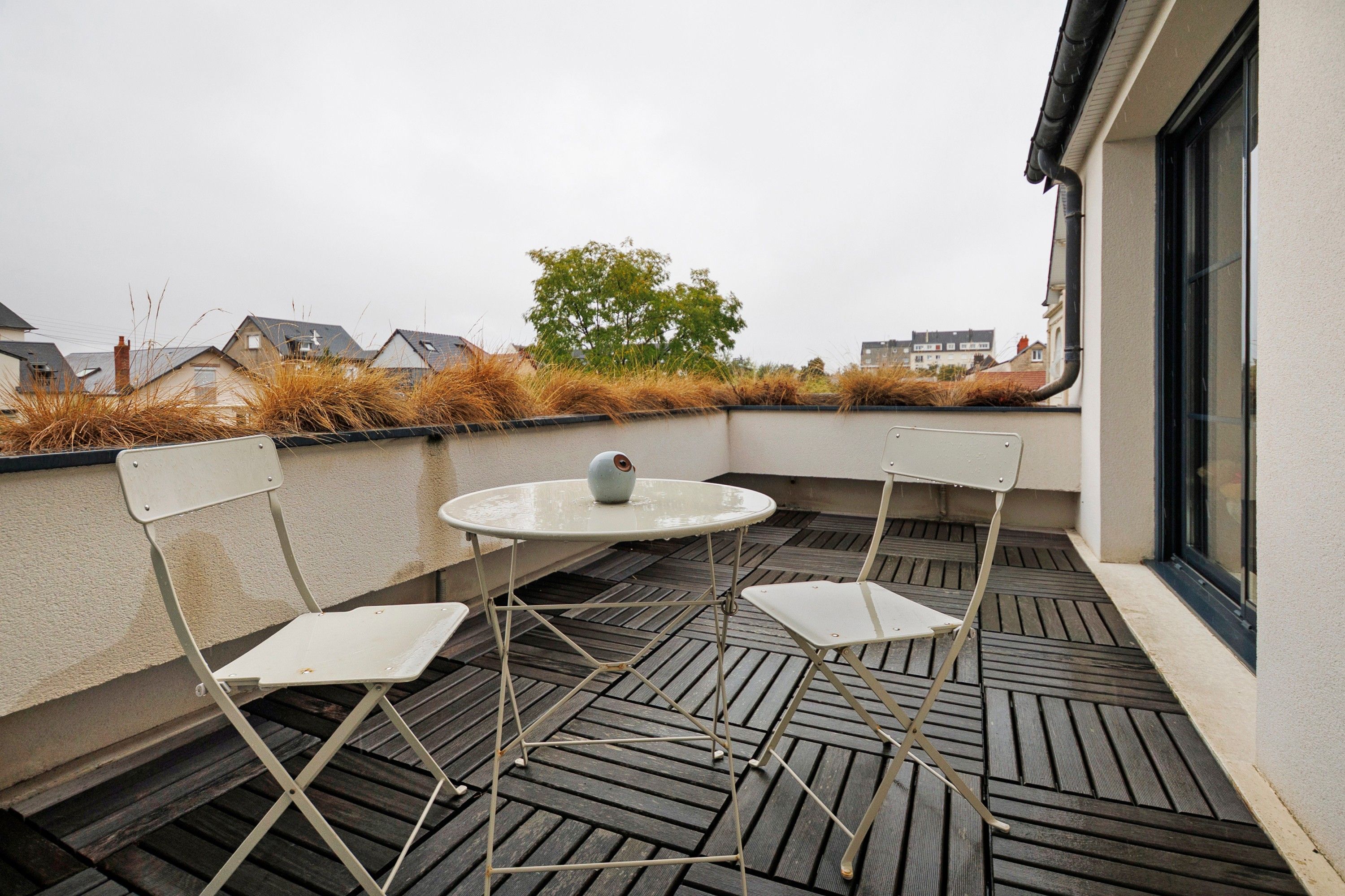 Une terrasse sur le toit, avec une table ronde et quatre chaises, un pot de fleurs et une vue sur les bâtiments voisins.