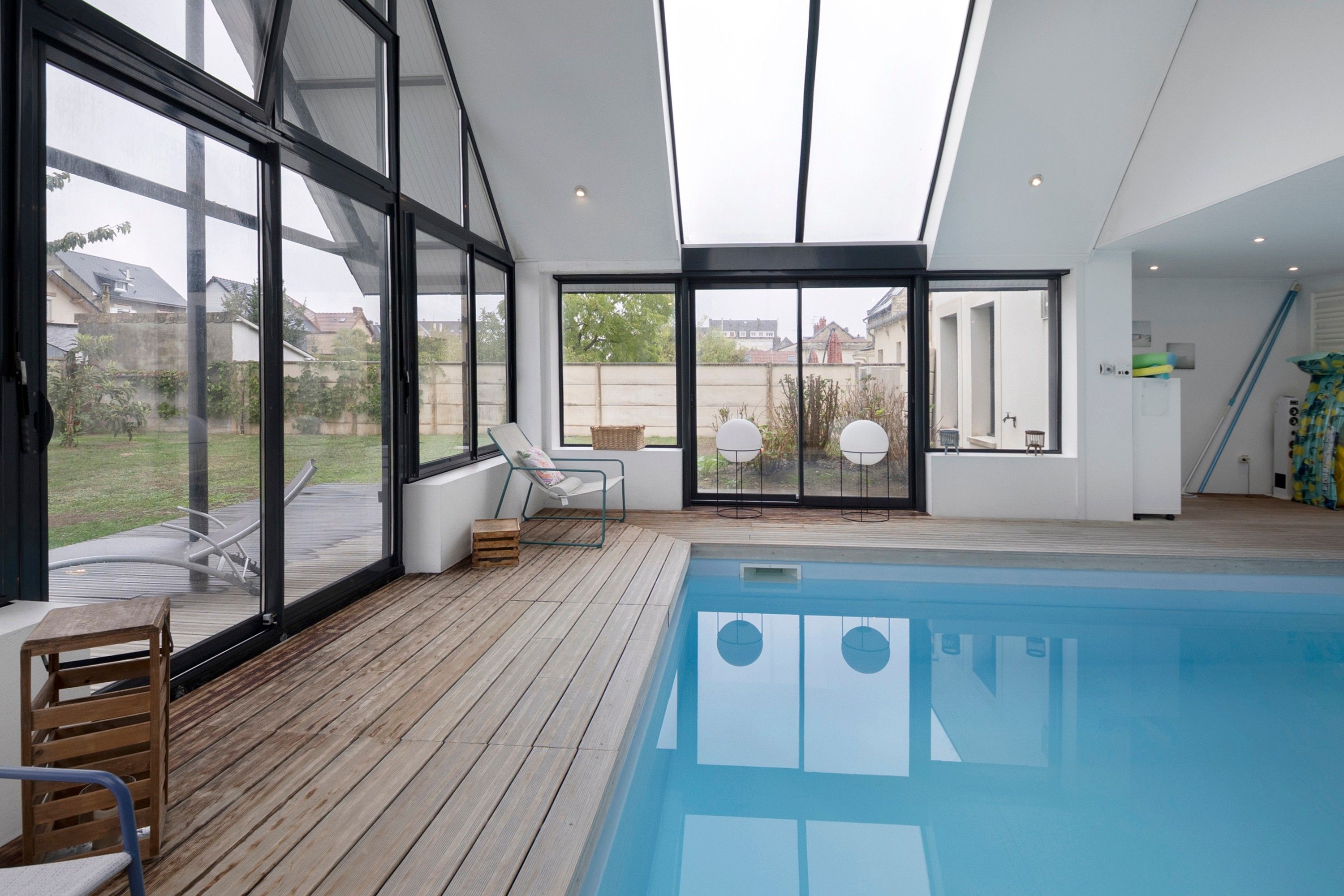 Une piscine intérieure moderne, dotée d’une terrasse en bois, de grandes fenêtres et offrant une vue sur les environs.