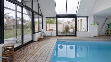 Une piscine intérieure moderne, dotée d’une terrasse en bois, de grandes fenêtres et offrant une vue sur les environs.