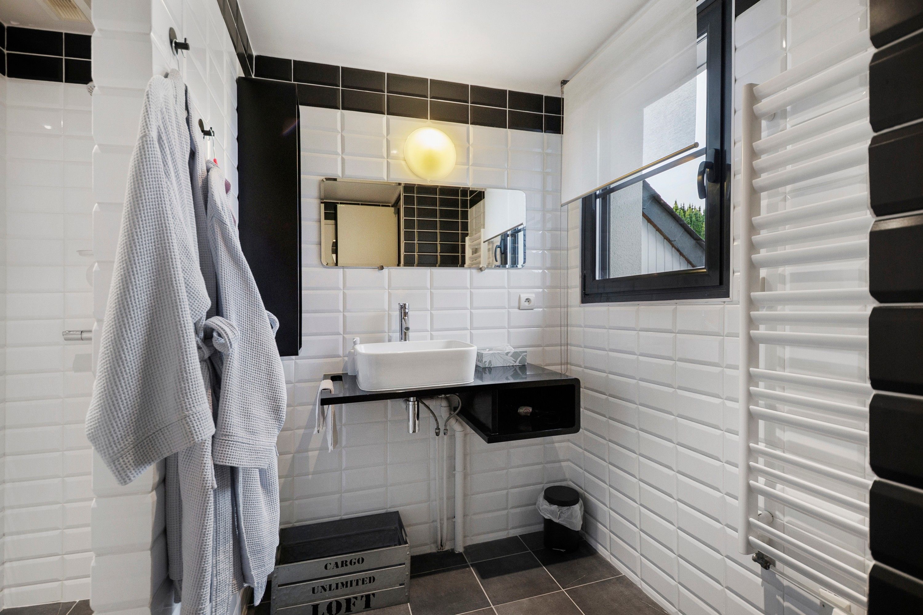 Un baño moderno con pared de azulejos blancos, tocador con lavamanos negro, espejo con luz redonda y ventana con vista a la vegetación.
