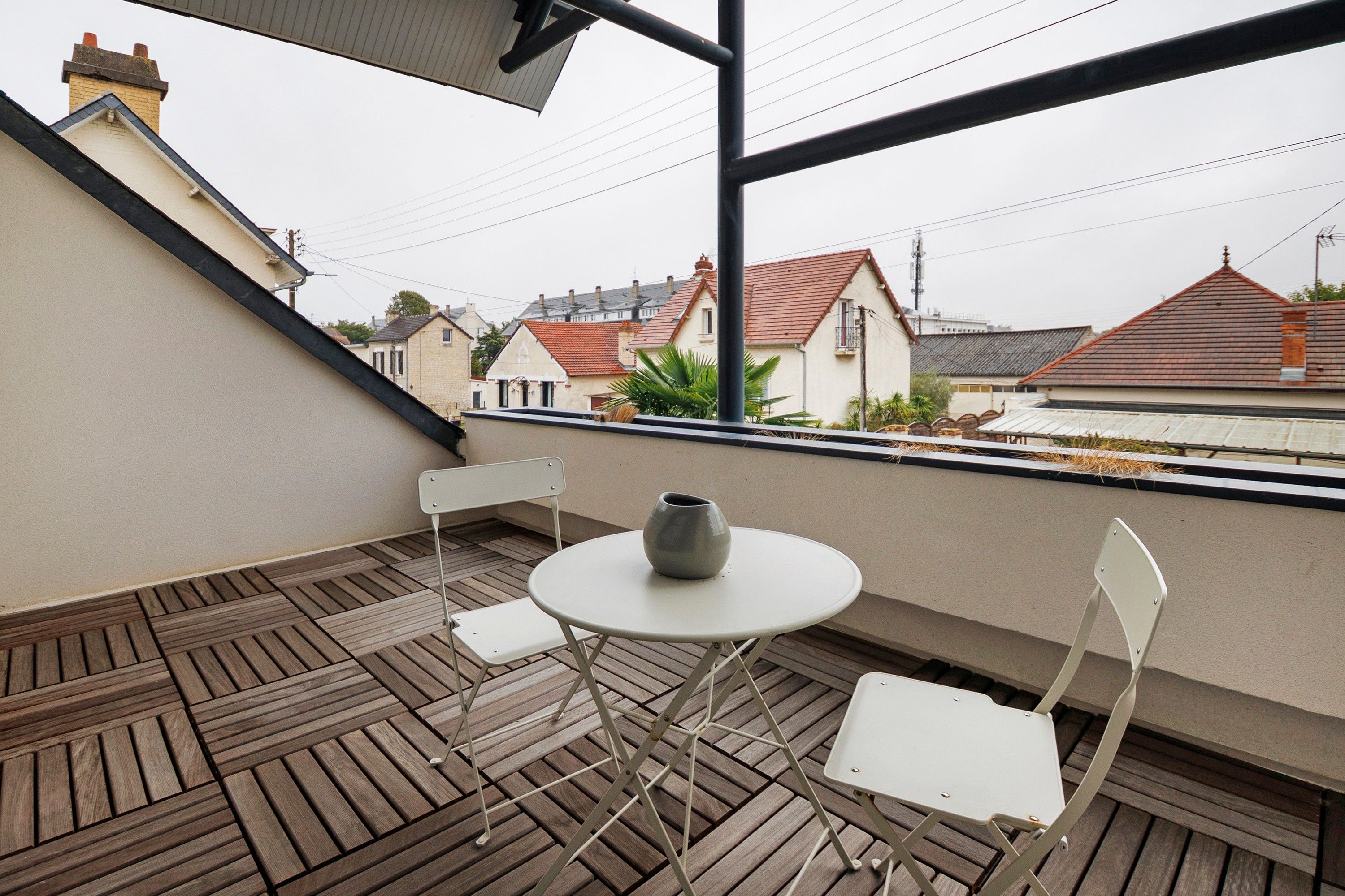 Un balcon avec une table ronde et deux chaises, donnant sur un quartier résidentiel avec des maisons.