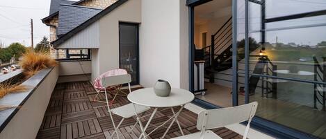 En udendørs terrasse med et bord, stole, en potteplante og udsigt til indretningen gennem en glasdør.