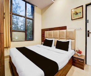 A bedroom with a large window, a bed with white and black bedding, a wooden headboard, and a small bedside table with a vase of flowers. - Classic Hotel Metro View (Jaipur)