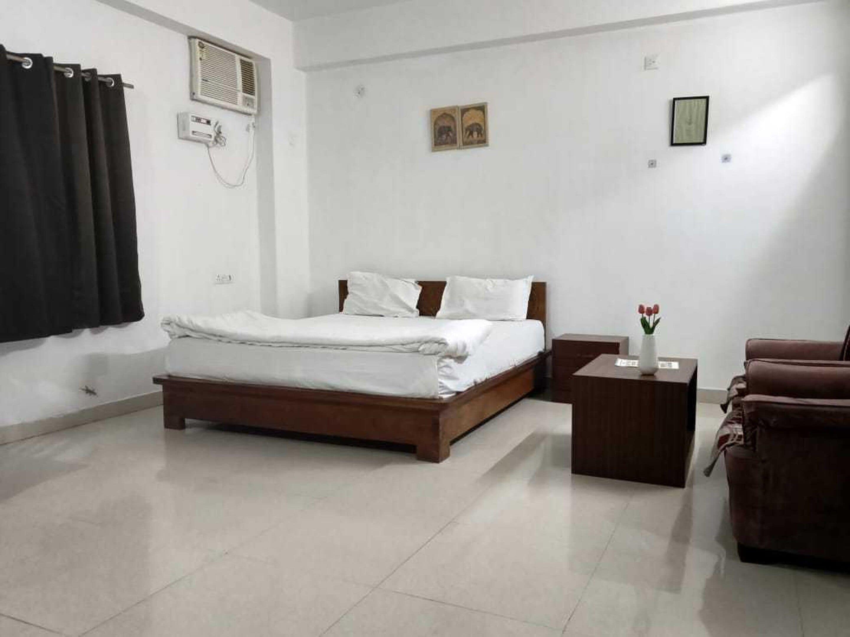 A hotel room with a bed, a nightstand, a chair, and a small table with a vase of flowers.