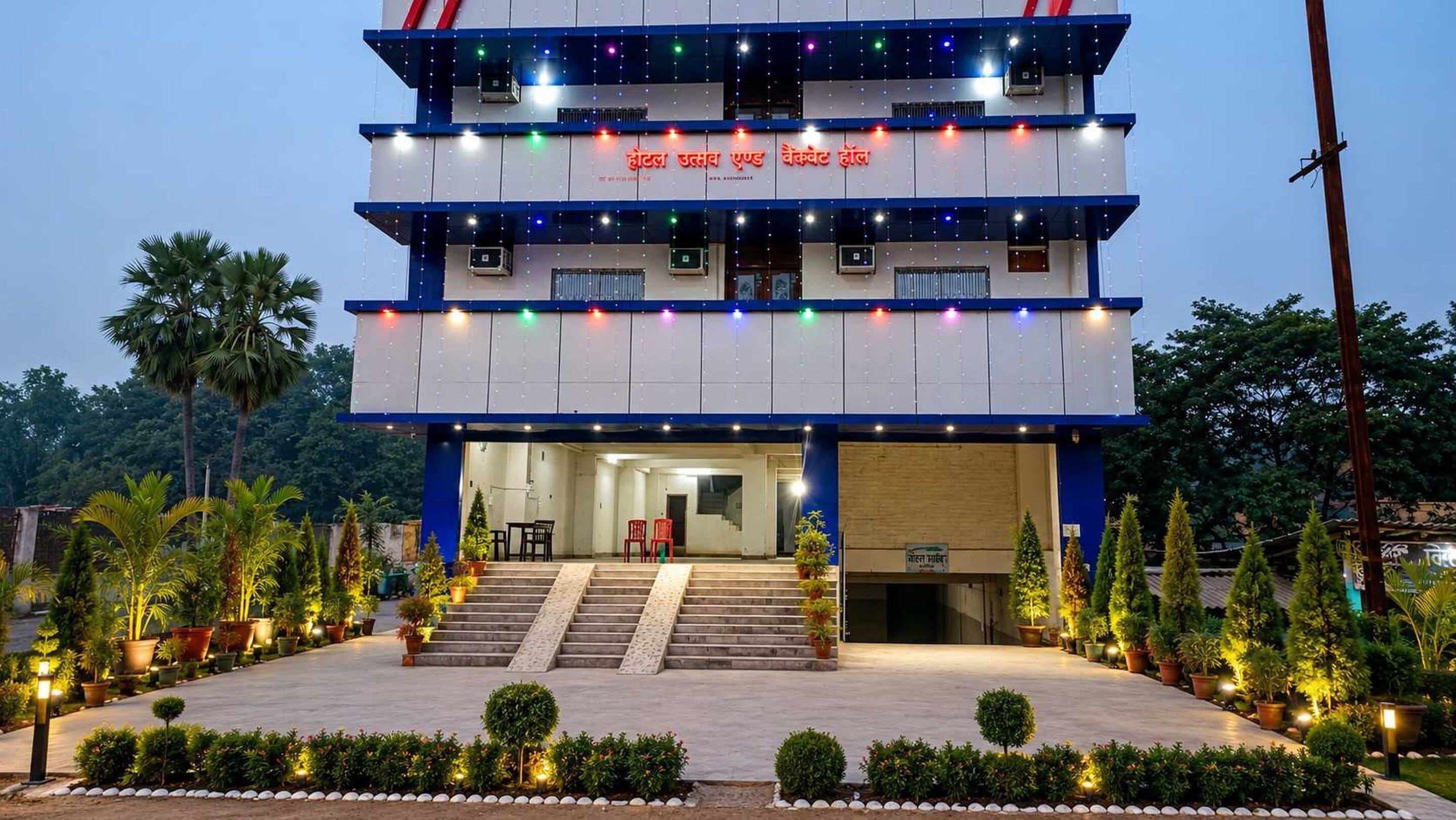 A multi-story hotel with a staircase leading to the entrance, surrounded by potted plants and a well-lit pathway.