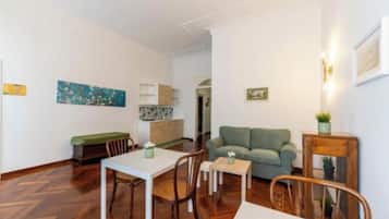 A living room with a sofa, a dining table with chairs, a small table with a plant, and a kitchen area in the background.