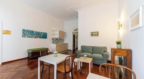 A living room with a sofa, a dining table with chairs, a small table with a plant, and a kitchen area in the background. - Marina Rooms (Napoli)