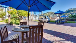 Un espace patio avec une table ronde en bois et des chaises, un grand parasol bleu et un camion garé au fond.